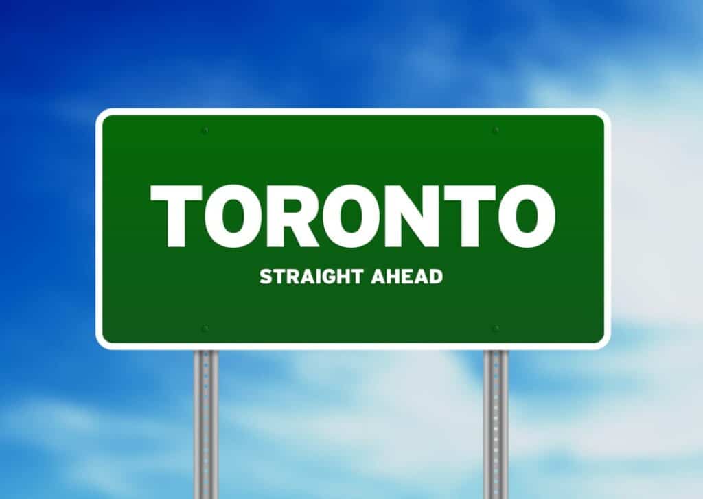 A green road sign against a blue sky, adorned with white clouds, reads "TORONTO" in bold white letters, with "STRAIGHT AHEAD" below in smaller text. Mounted on two metal poles, it's the perfect guide for travelers heading to car rentals or other adventures in Toronto.
