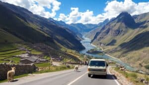 A white van drives down a winding mountain road in Peru's Sacred Valley, beside a scenic river. Sheep stand near the road, terraced houses dot the hillside, and tall green mountains rise beneath a bright blue sky filled with scattered clouds.