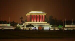 The Ho Chi Minh Mausoleum in Hanoi, Vietnam, is illuminated at night with warm golden and red lights. The large stone building draws visitors who often drive from afar, including from the vibrant city of Ho Chi Minh City or the lush Mekong Delta.