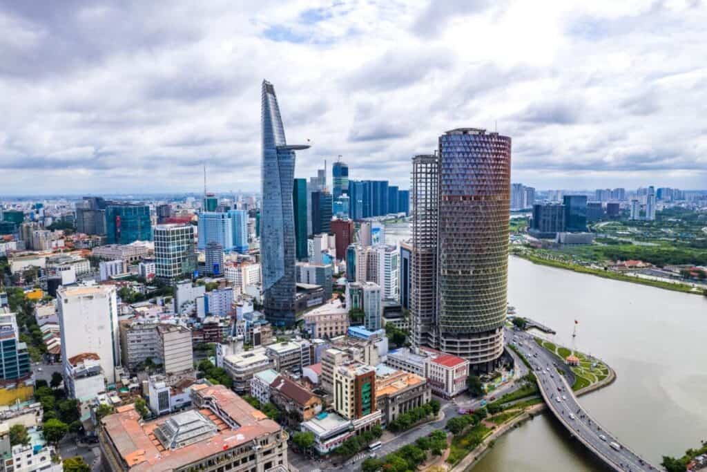 Aerial view of Ho Chi Minh City with tall skyscrapers, including a unique, pointed tower and a cylindrical building, near the river and bridge. The cityscape highlights dense buildings, greenery, the Mekong Delta nearby, and a cloudy sky above.