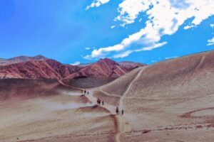 A group of people hikes along a winding trail through reddish-brown desert hills in China under a bright blue sky. Distant mountains and sandy terrain evoke the historic landscapes of the ancient Silk Road.