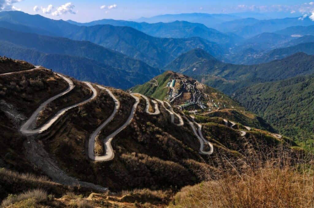 A winding mountain road with numerous sharp hairpin bends snakes up a steep, rugged hillside—a perfect scene for an epic journey through lush green landscapes, rolling hills, and distant blue mountains under a partly cloudy sky.