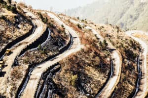 A winding mountain road, ideal for an epic journey or China road trip, twists through rugged terrain with sharp switchbacks. Construction vehicles dot the route, while distant hills and sparse vegetation fade into a misty backdrop.