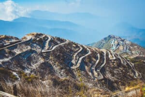 A winding mountain road with sharp hairpin bends cuts across dry, brown hills. Patches of snow and layers of blue mountains evoke the spirit of an epic journey on a China road trip, fading into the misty distance beneath a clear, slightly cloudy sky.