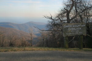 A wooden sign reads "Onion Mountain Overlook, Elev 3195, 6 minute loop trail" beside a road on the Blue Ridge Parkway. Bare trees frame blue-tinged mountains under a hazy sky, offering a scenic trip overview for travelers. Grass lines the roadside.