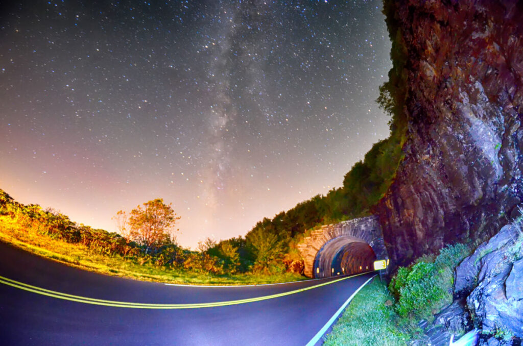 A curving, empty road on the Blue Ridge Parkway leads to a stone tunnel carved into a rocky cliff at night. The sky sparkles with bright stars and the Milky Way, as roadside plants and rocks glow under colorful lights.