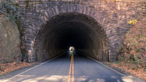 A paved road with yellow center lines leads into a dark, stone-arched tunnel through a rocky hillside on the Blue Ridge Parkway. Sunlight glows at the tunnel’s end, and fallen leaves are scattered along the road’s edges—an essential guide for your trip overview.