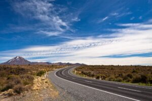 A winding road curves through a landscape of brown scrub on New Zealand’s North Island, with a snow-capped mountain in the distance beneath a blue sky streaked with wispy clouds—perfect for a scenic road trip.