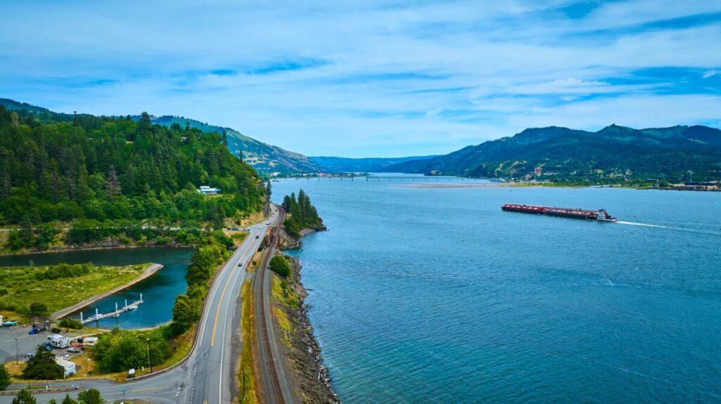A wide river runs between forested hills under a blue sky. The timeless Historic Columbia River Highway curves along the left riverbank, with boats docked at a small marina as a long barge travels toward a distant bridge connecting both shores.