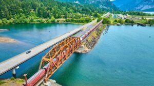 Aerial view of a rust-colored steel train bridge and the Historic Columbia River Highway, both crossing a wide river surrounded by lush trees and mountains in the background, with cars on the road and a train on the bridge.