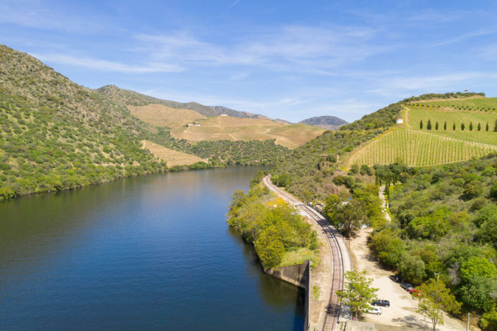 A wide river curves between green, hilly landscapes in the Douro Valley, with vineyards on the right. A railway track runs parallel to the river along tree-lined banks. Several parked cars mark scenic Vineyard Stops. The sky is clear with a few wispy clouds.