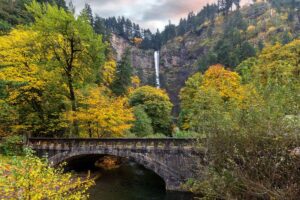A stone bridge crosses a stream surrounded by trees with green and autumn yellow leaves. In the background, a tall waterfall cascades down a forested cliff under a cloudy sky along the Historic Columbia River Highway.