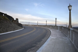 Curved road and sidewalk bordered by a stone wall with vintage-style street lamps; reminiscent of the timeless Historic Columbia River Highway, stone stairs rise at left, sky above is pale blue with sparse clouds, scene empty of people or vehicles.