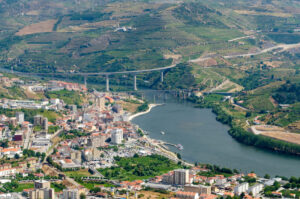 Aerial view of a river curving through a city with clusters of buildings in the foreground, green hills and farmlands in the background, and a long modern bridge spanning the Douro Valley amid vineyard stops along this scenic wine drive.