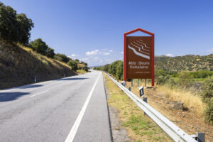 A rural road winds through hilly terrain with trees and dry grass. On the right, a red sign marks "Alto Douro Vinhateiro," welcoming you to Portugal's UNESCO World Heritage Douro Valley—perfect for scenic vineyard stops on your wine drive.