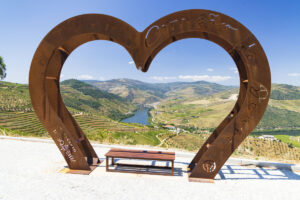 A large, rust-colored metal heart sculpture frames a scenic view of terraced vineyards and rolling hills—an ideal stop on a wine drive with vineyard stops, inviting visitors to relax and soak in the picturesque Douro Valley landscape from a wooden bench.