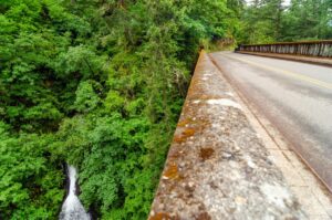 A road and bridge, part of the Historic Columbia River Highway, run alongside a dense green forest. Weathered railings line the bridge, while trees cover steep cliffs below. A small waterfall peeks through thick greenery at the bottom left.