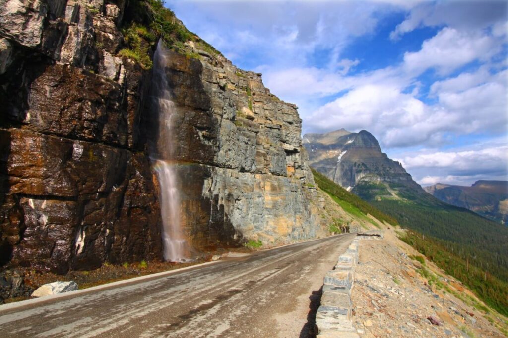 A narrow waterfall cascades down a rocky cliff beside the famous Going-to-the-Sun Road in Glacier National Park, USA. Wet pavement reflects sunlight as it meanders through this stunning landscape. In the background, green forest and tall, rugged mountains rise under a partly cloudy blue sky. The scene is bright and peaceful.