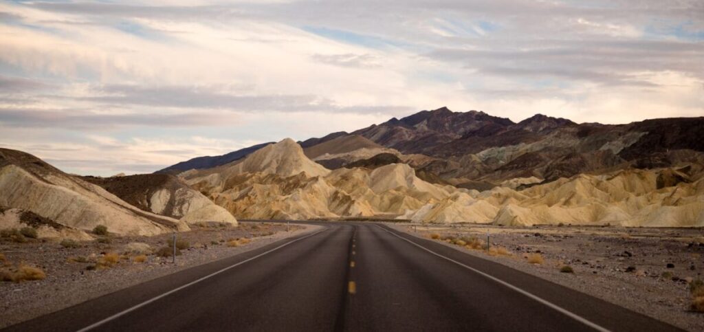 A straight, empty road stretches into the distance toward rugged, pale yellow and brown hills with a dark mountain backdrop under a partly cloudy sky in this striking Death Valley desert landscape. Sparse vegetation lines the roadside, making it an ideal route for a scenic drive or as a photographic guide to the region’s natural beauty.
