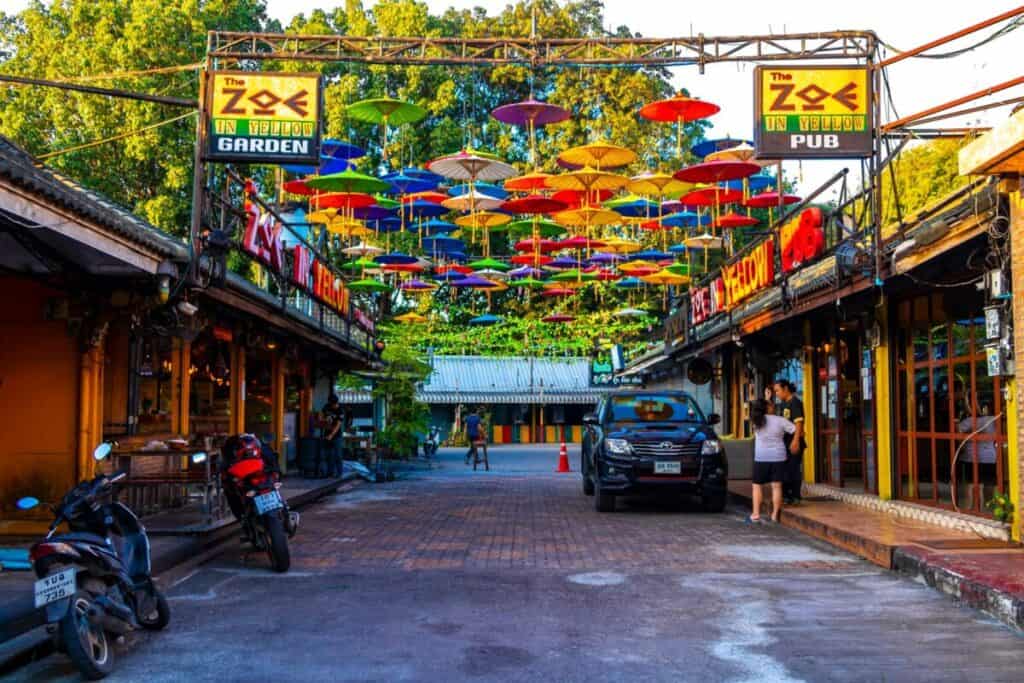 A lively street in Chiang Mai, Thailand, lined with bars and restaurants, displays colorful umbrellas hanging overhead. Motorbikes and a black car are parked nearby, and two people stand in front of a building. Signs read “Zoe in Yellow Pub” and “Garden.” Trees fill the background along this vibrant stop on the Northern Loop.