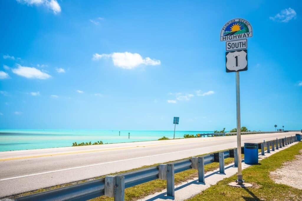 A sunny coastal highway with clear turquoise water on the left, a blue sky above, and a “Highway 1 South” road sign in the foreground offers the perfect backdrop for a scenic drive through the Florida Keys. There’s a blue trash bin and guardrail beside the road, and lush greenery along the water’s edge, inviting you to embrace adventure at every turn.
